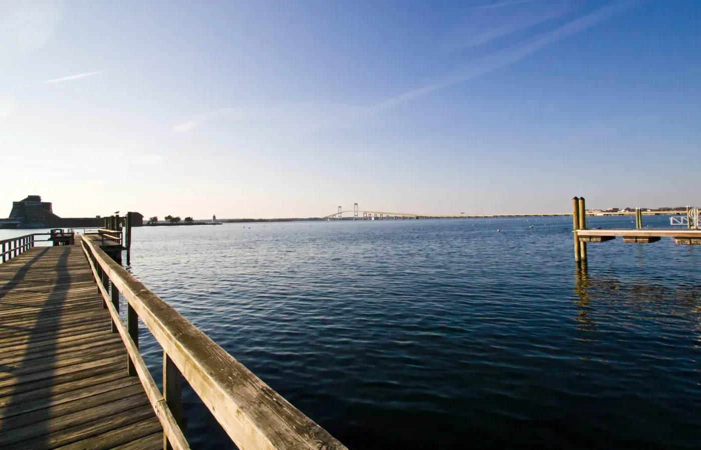 Wooden boardwalk overlooking calm blue water at sunset, with a distant city skyline under a clear sky