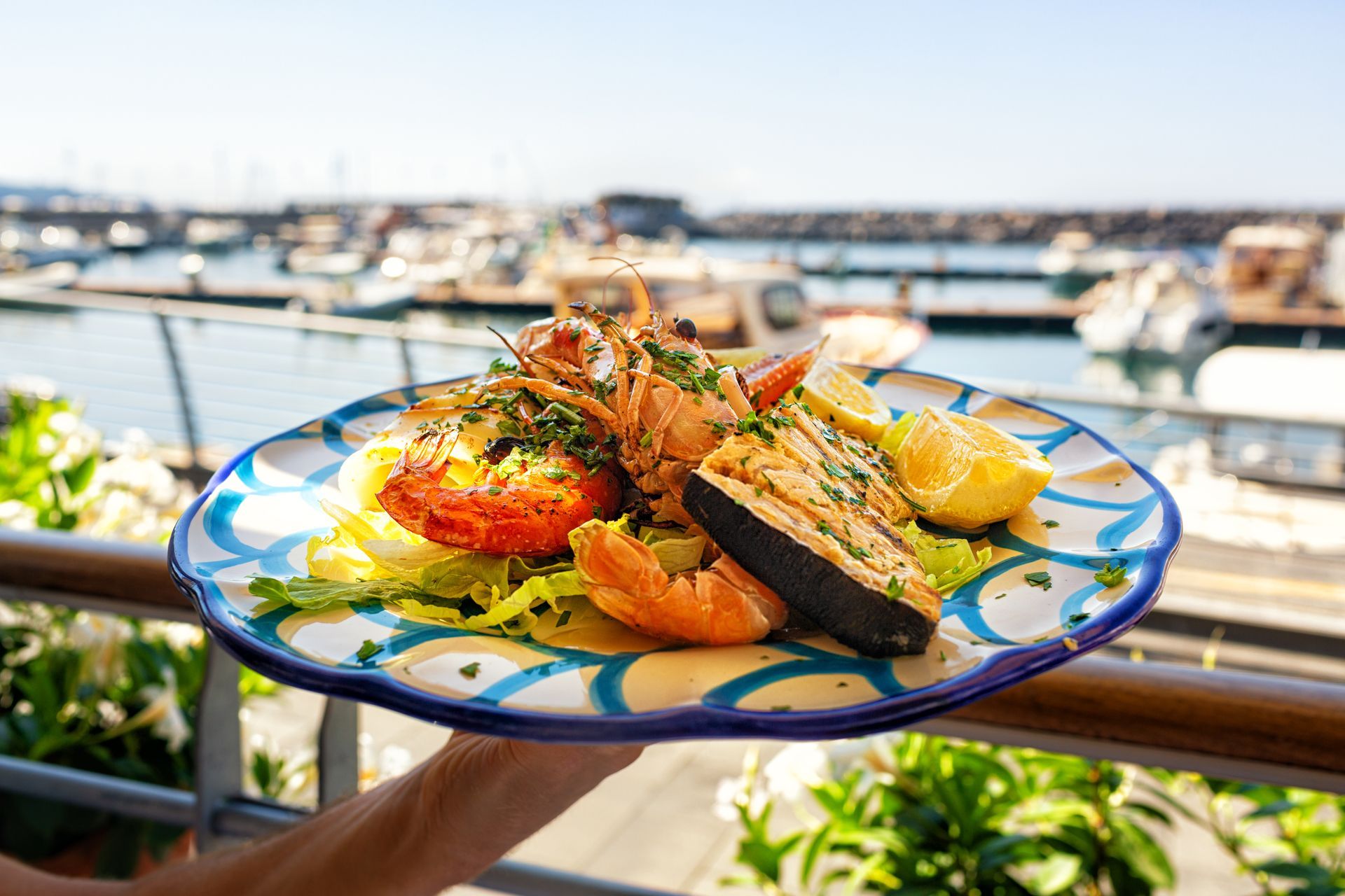 Colorful seafood dish on a blue-rimmed plate with boats in a sunny marina background