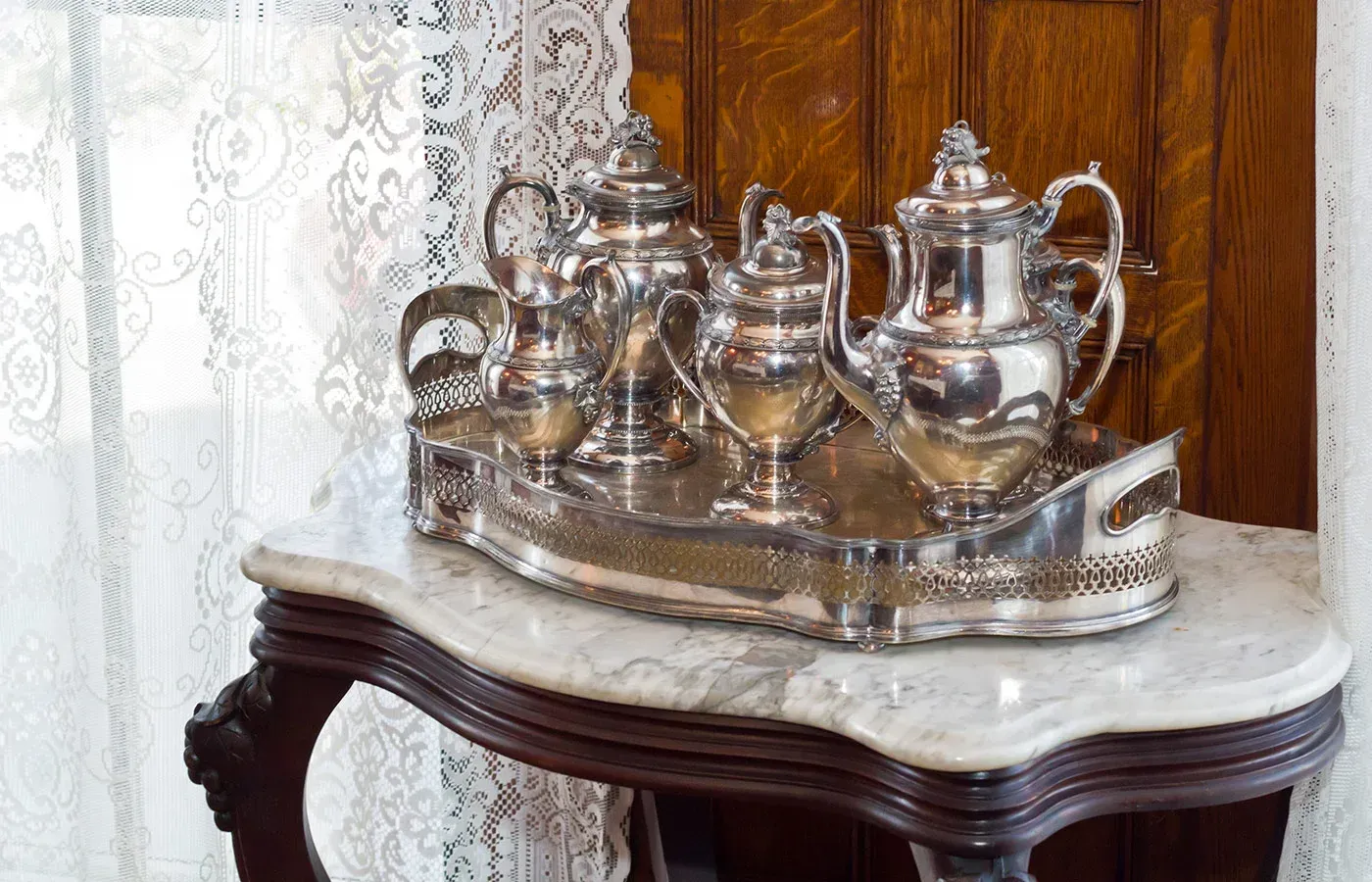 Silver tea set on a tray atop a marble-topped wooden table by a lace curtain
