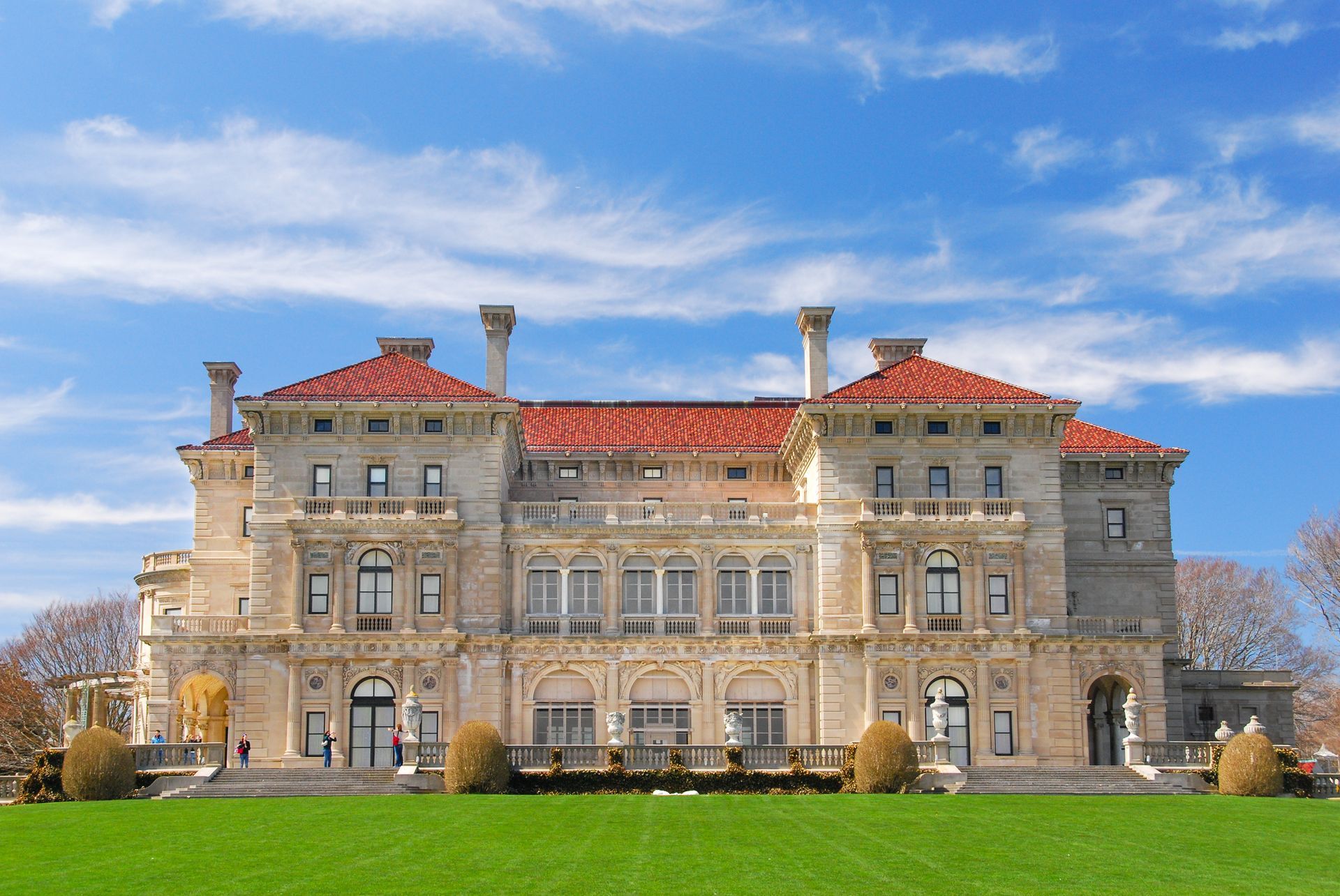 Large stone mansion with red roof on a green lawn under a blue sky