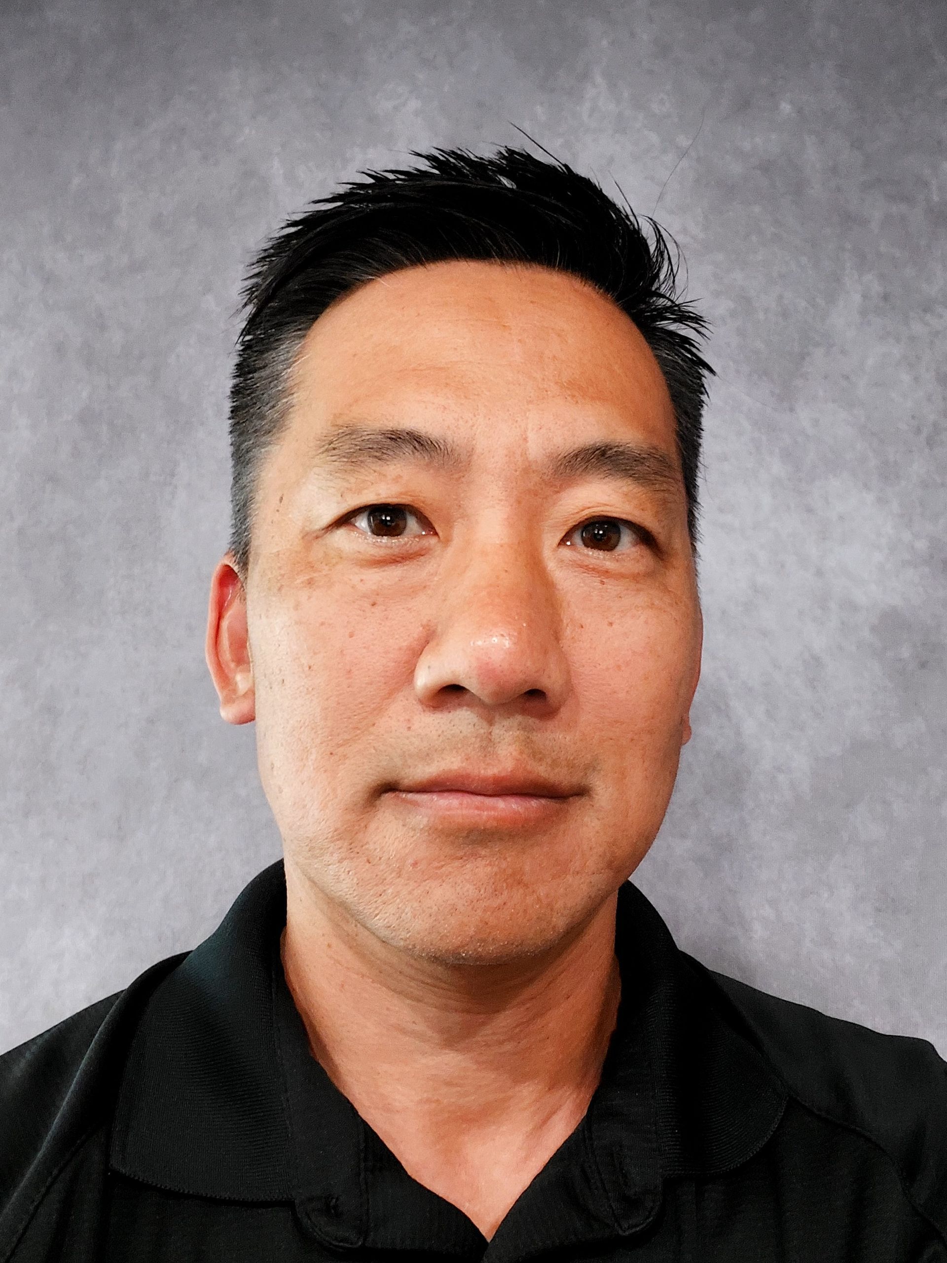 Man with short black hair, wearing a black shirt, against a gray background, looking forward. | AK Auto Repair