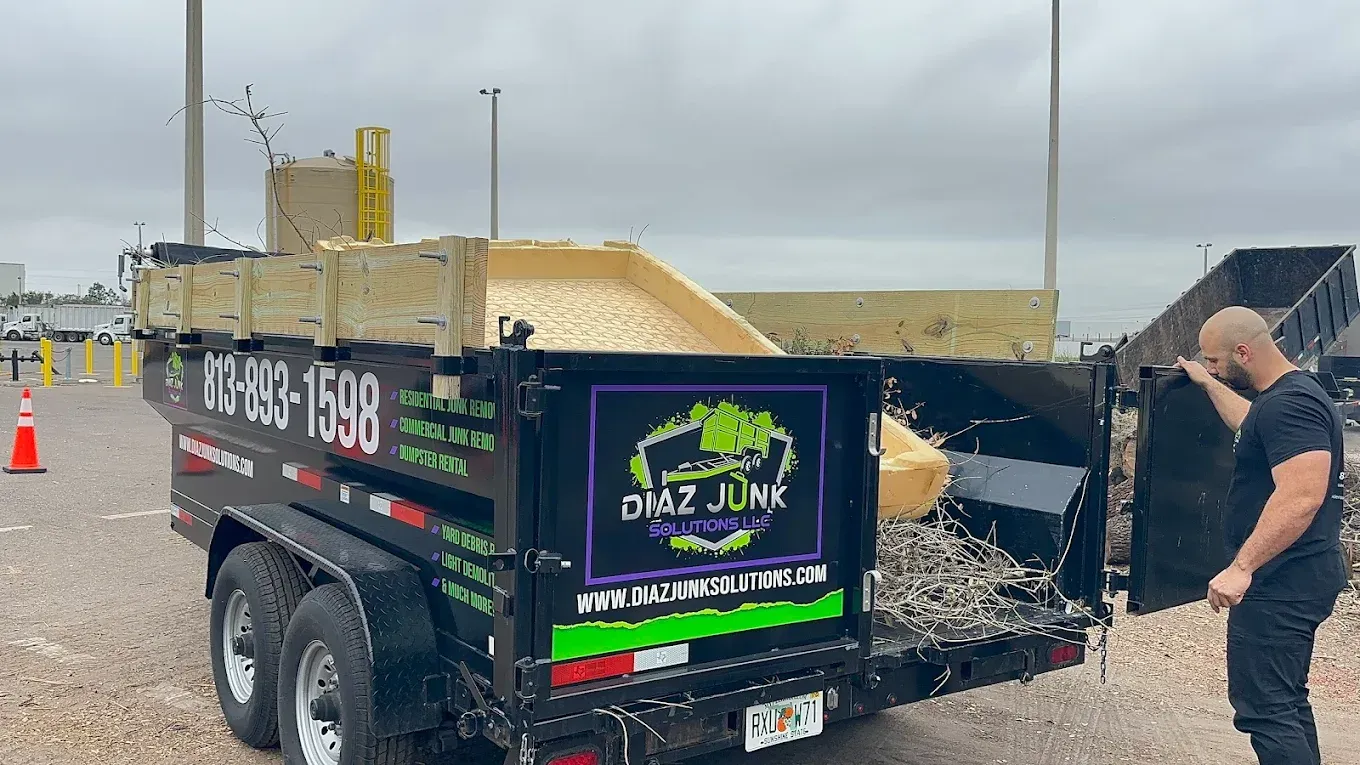 A man unloading debris from a black trailer with “DMZ Junk” logo. Cloudy outdoor setting.