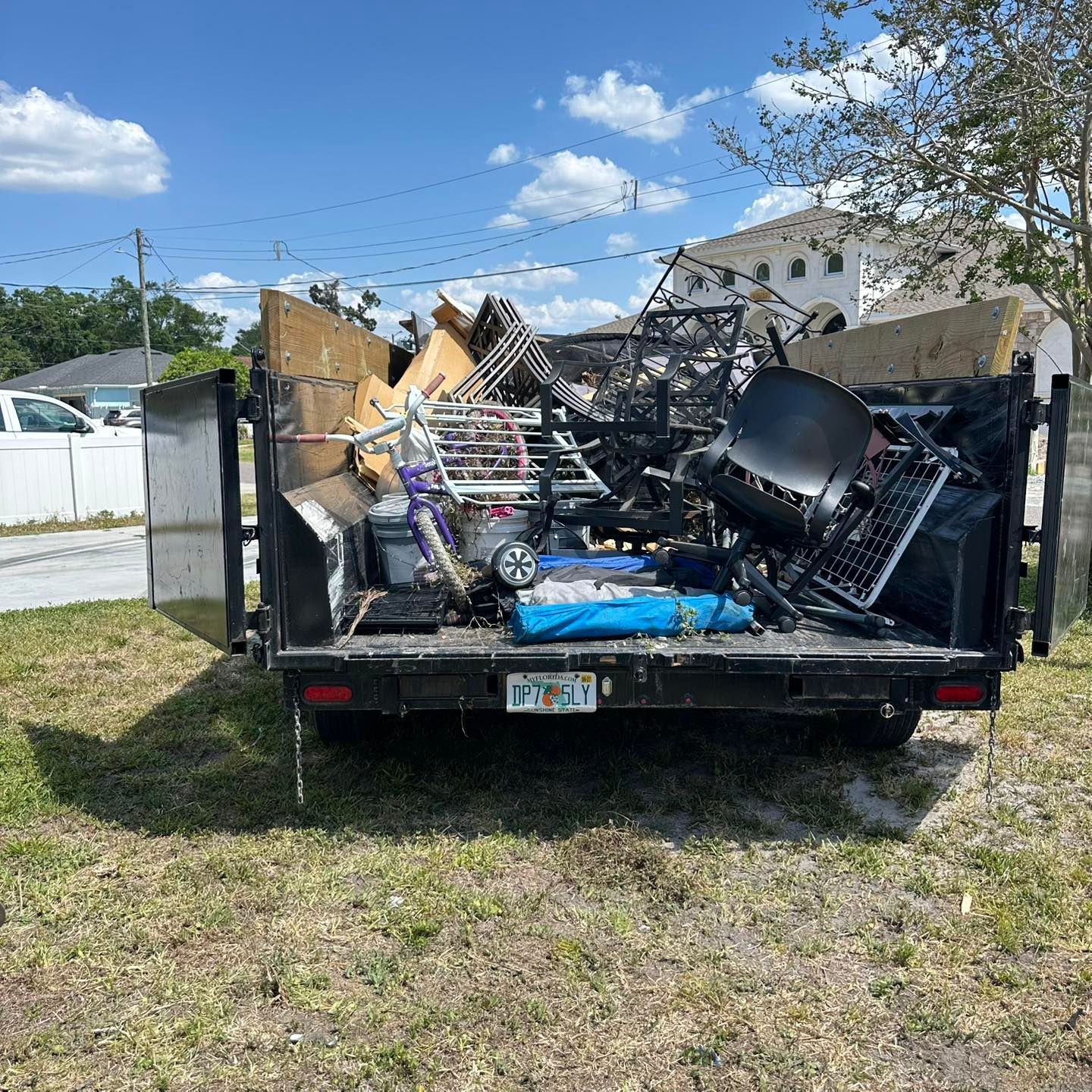 A black trailer overflowing with various debris, including furniture and metal, parked on a grassy area under a blue sky.