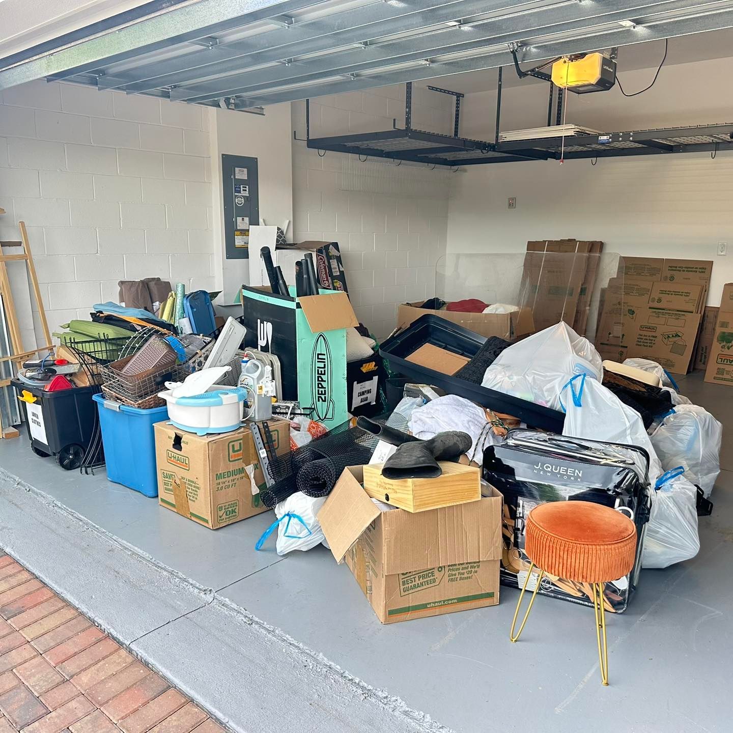 Cluttered garage with boxes, bags, and various items. An orange stool sits in the front.