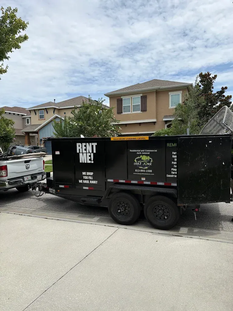 Black rent-me trailer parked on a suburban street beside a pickup truck and houses.