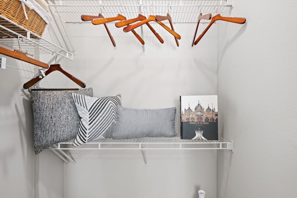 A neat and organized closet with white wire shelves. On the shelves, there are several decorative pillows with different textures and patterns, a wicker basket with a white linen sticking out, and a hardcover book titled Wonders of the World propped up against the wall. Above, wooden hangers hang on the rack, some holding nothing and others with hangers attached to them. The closet is well-lit, and the walls are painted white, giving the space a clean, minimalist appearance at Marq Inverness in Englewood, CO.