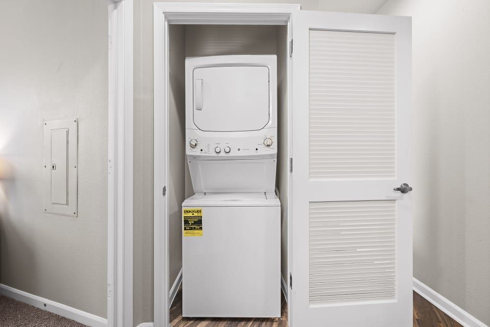 A stackable white washer and dryer set enclosed in a small laundry closet with bifold doors, one of which is open, set against neutral-colored walls in a residential interior at Marq Inverness in Englewood, CO.