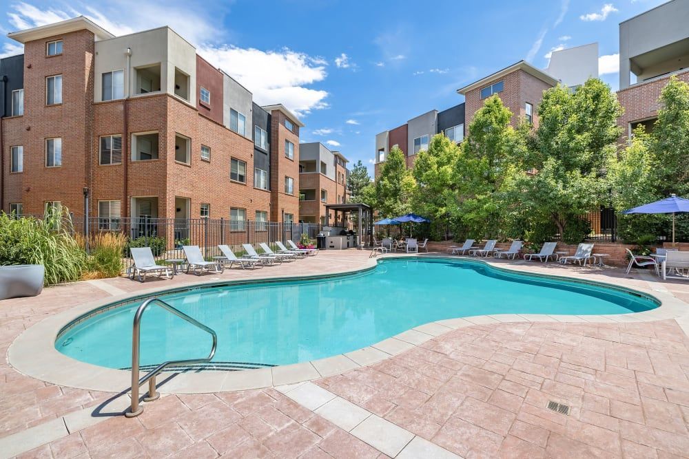 Outdoor swimming pool at a modern apartment complex, surrounded by lounge chairs and shaded tables with umbrellas, flanked by multi-story brick apartment buildings with lush green trees at Marq Inverness in Englewood, CO.