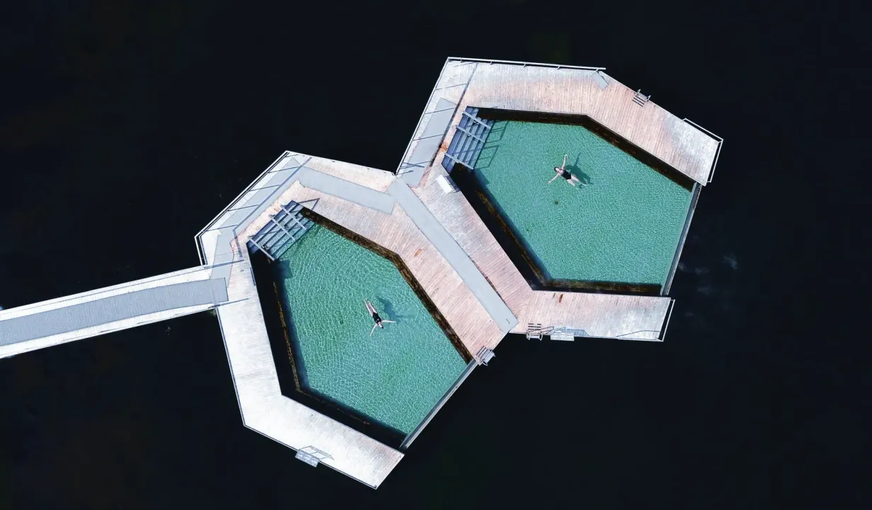 Aerial view of two connected hexagonal turquoise structures on dark water with a narrow walkway.