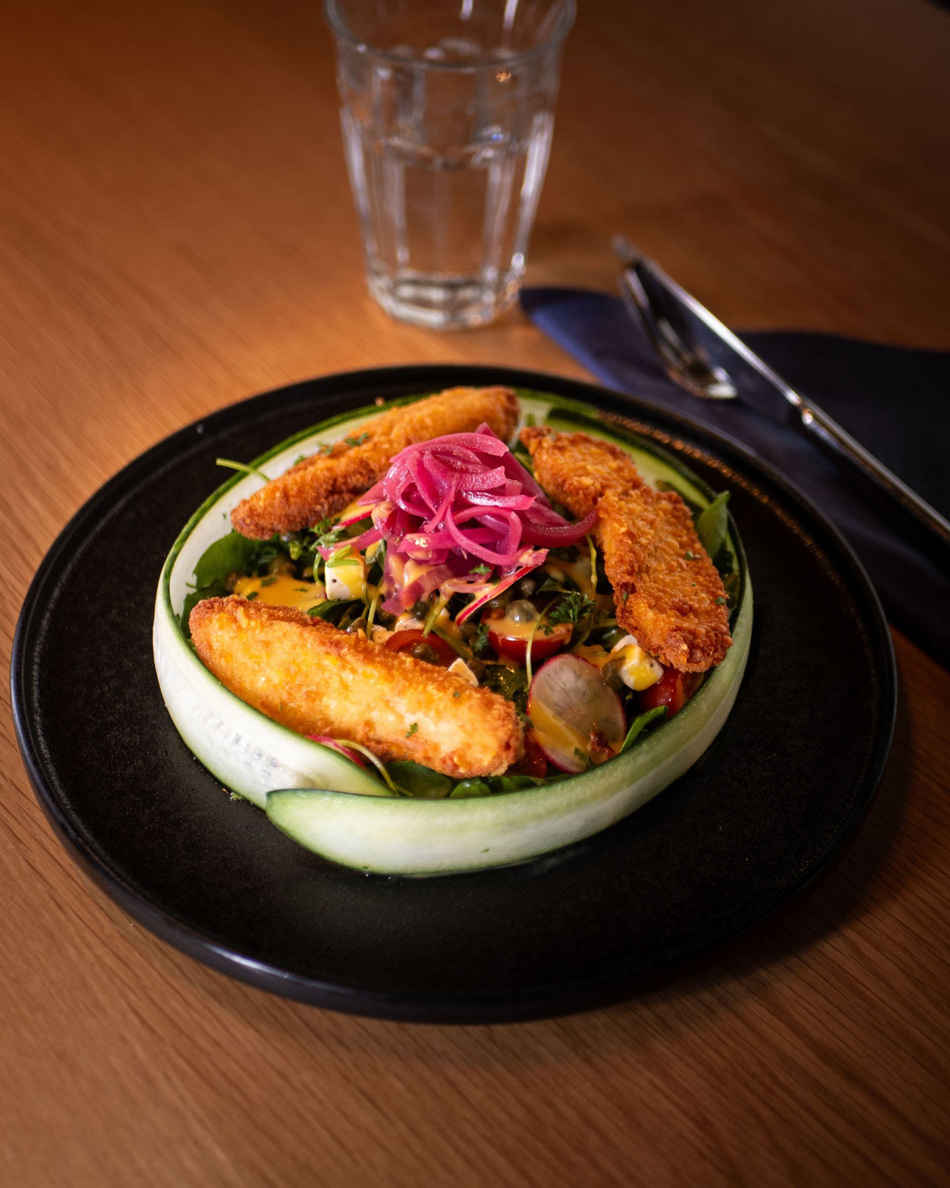 Plated gourmet dish with fried shrimp, sliced cucumber, and pink pickled onions on a black plate