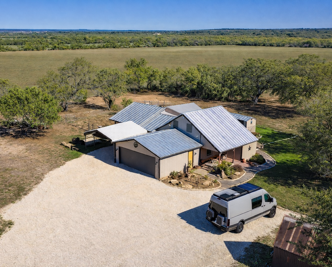 Rural homestead with metal-roofed buildings, gravel yard, and a parked SUV in open countryside