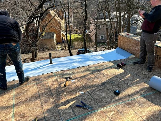 Two people installing blue underlayment on a roof in a wooded backyard