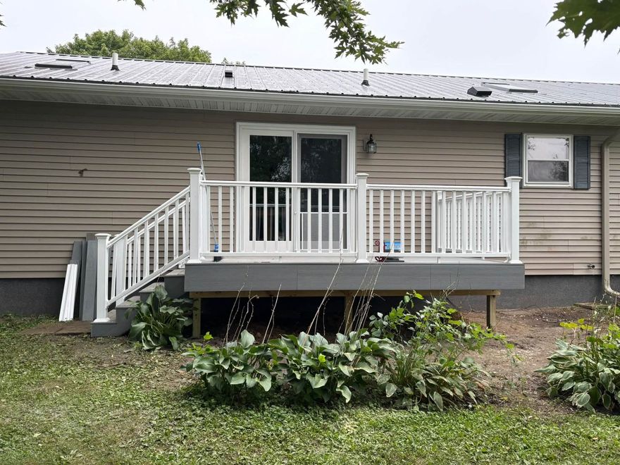 Small beige house with white porch and railing, seen from the yard, with shrubs and trees around it.