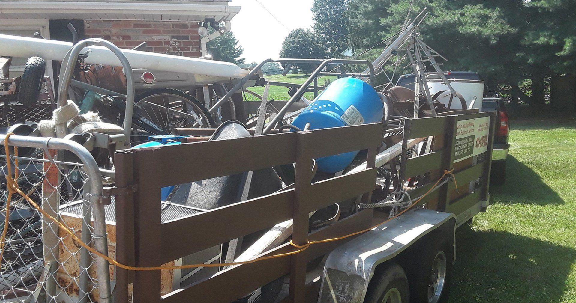 Utility trailer loaded with metal frames and blue items, parked on a grassy yard beside a house.