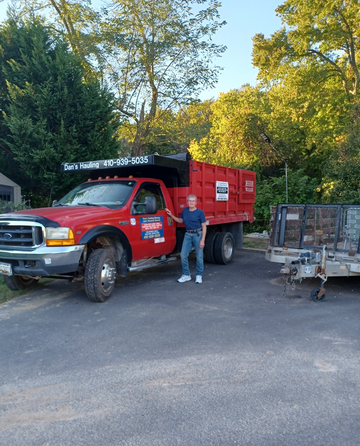 Red dump truck parked on a gravel road with a person standing beside it near a trailer and trees.