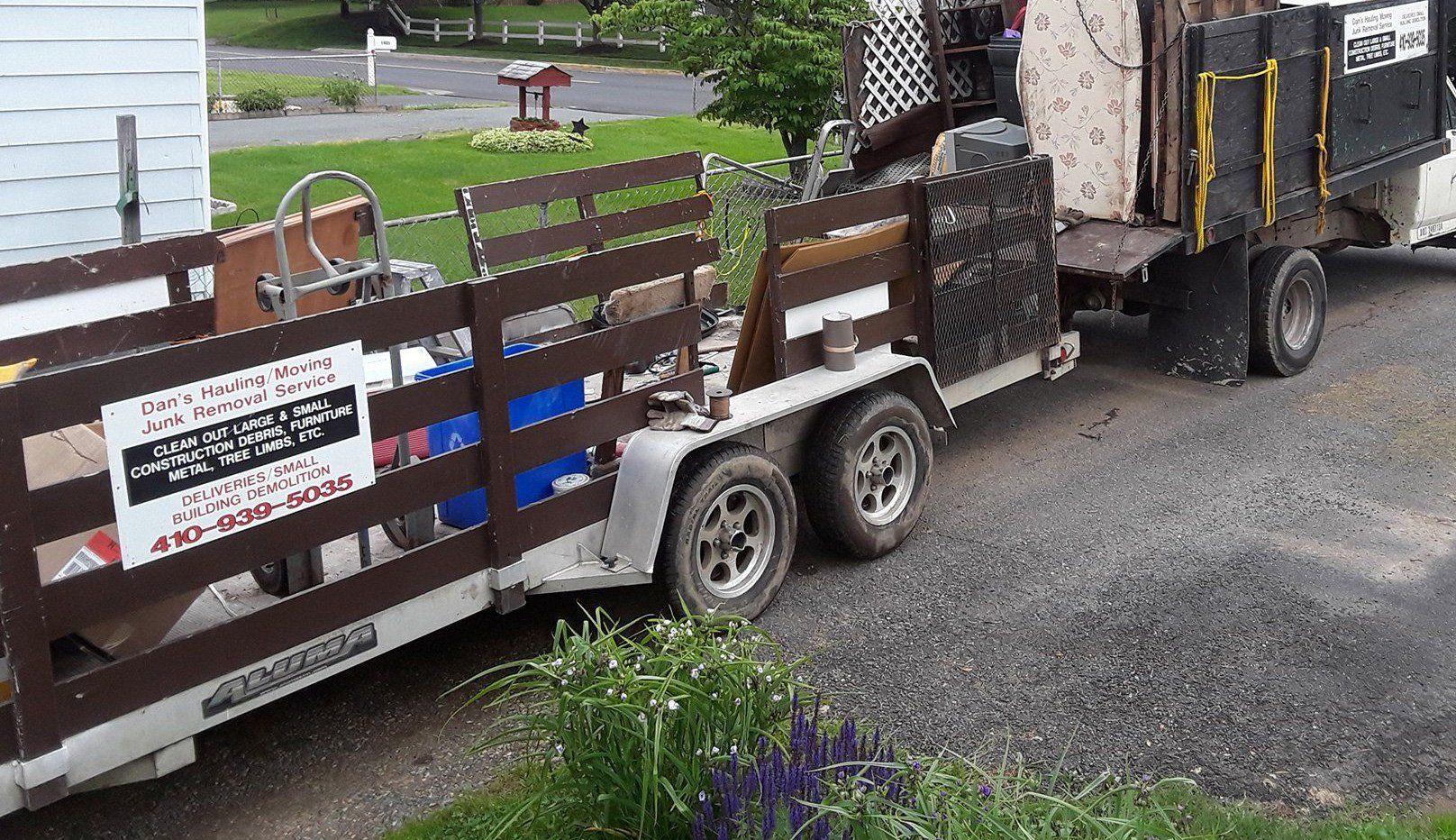 Small trailer with wooden sides carrying a large machine, parked on a gravel driveway beside a house.