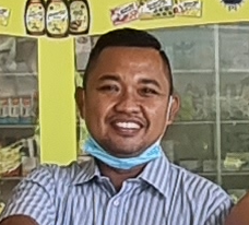 Seorang pria yang mengenakan masker wajah tersenyum ke arah kamera, di dalam sebuah toko dengan rak-rak berisi produk.