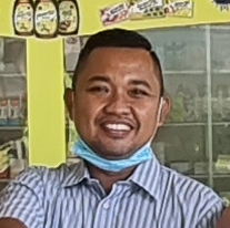 Pria mengenakan kemeja bergaris dan masker wajah tersenyum di depan rak di sebuah toko.