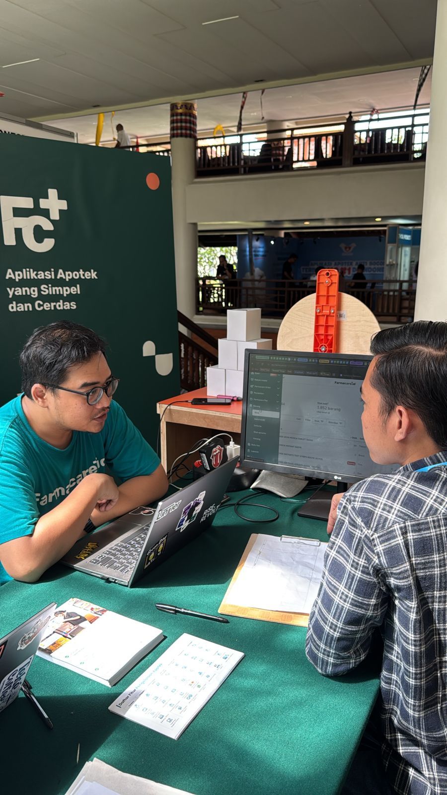 Dua pria di meja, satu menunjukkan layar laptop kepada yang lain. Latar belakang hijau dengan logo, kotak-kotak lain, dan monitor terlihat.