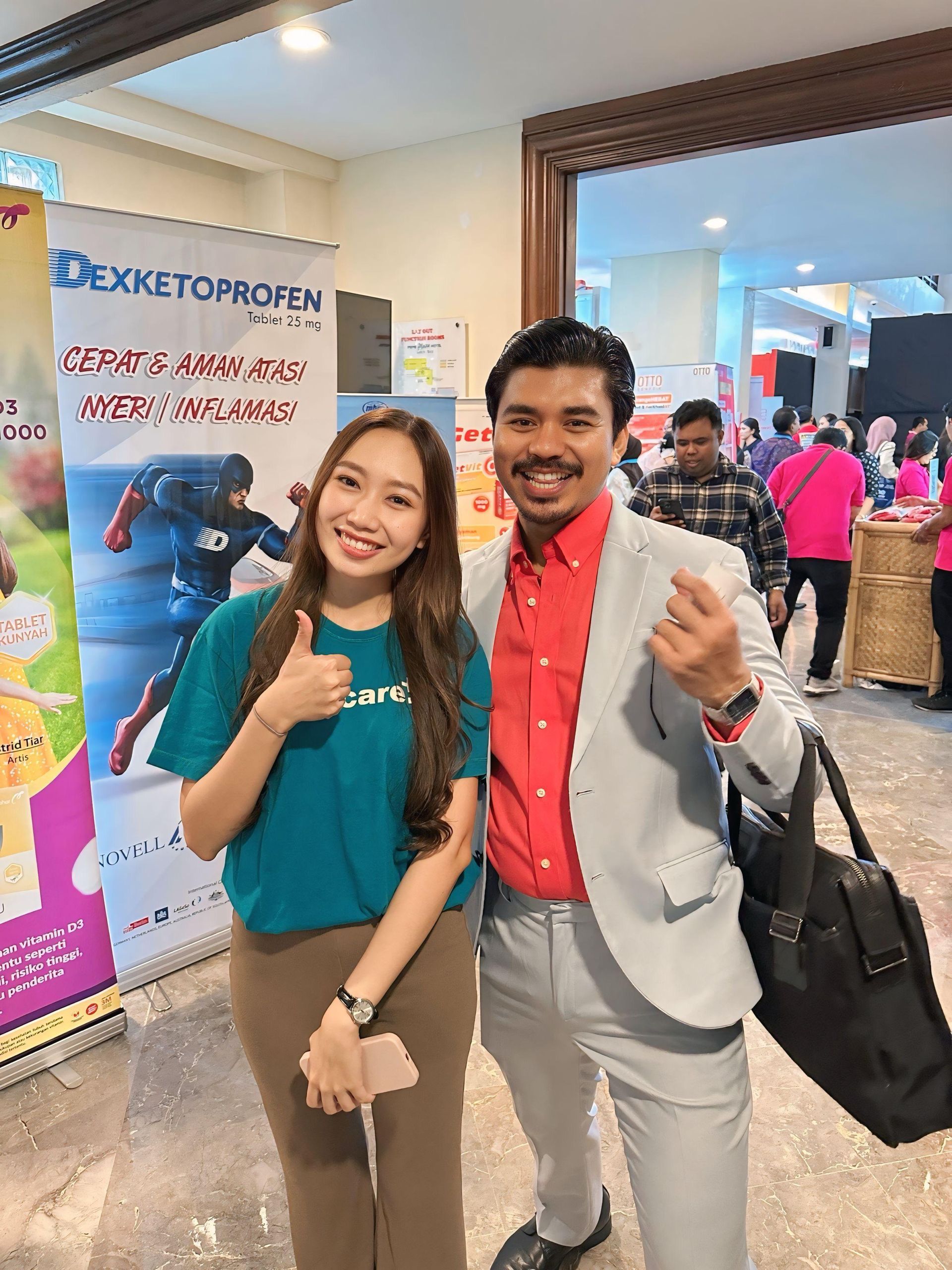 Wanita berbaju biru kehijauan mengacungkan jempol. Pria berjas abu-abu memberi tanda perdamaian. Mereka berdiri di dekat spanduk di dalam ruangan.