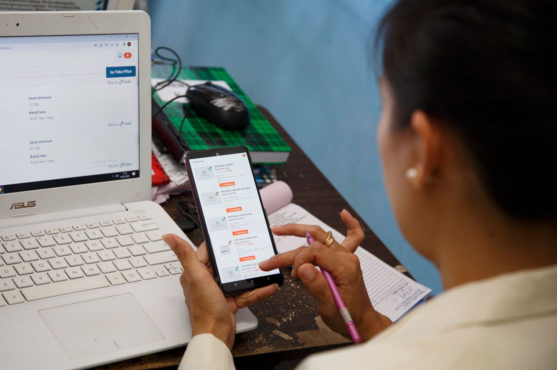 Seseorang menggunakan ponsel dan laptop di meja; ponsel menampilkan grafik. Mouse komputer dan buku catatan terlihat.