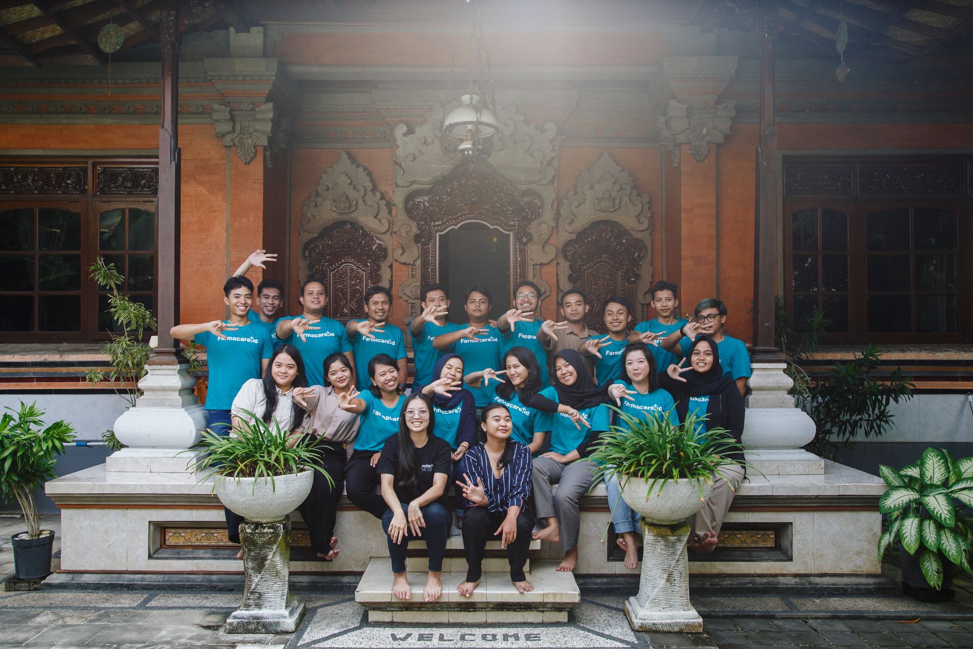 Sekelompok orang berbaju biru kehijauan berpose di tangga depan sebuah gedung dengan ukiran hiasan, beberapa membuat gestur.