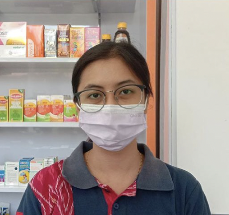 Seorang wanita berkacamata dan memakai masker berdiri di depan rak-rak produk.