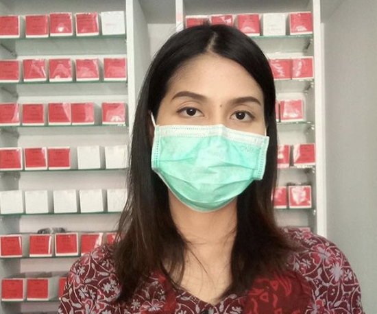 Seorang wanita mengenakan masker bedah di depan rak-rak berisi kotak-kotak merah dan putih.