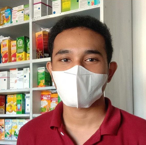 Pria bermasker, berbaju merah, di apotek, rak putih berisi obat-obatan di belakangnya.