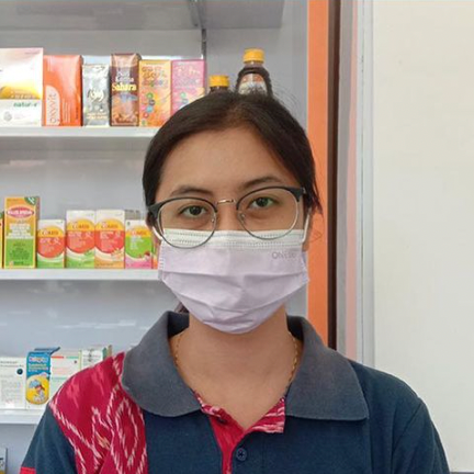 Wanita mengenakan kacamata dan masker wajah di apotek, rak produk di belakangnya.