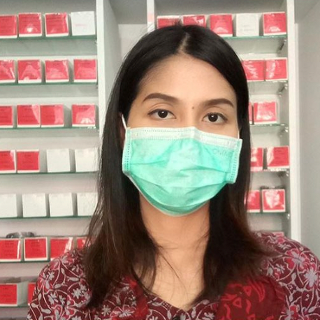 Wanita mengenakan masker bedah di depan rak dengan kotak putih dan merah.
