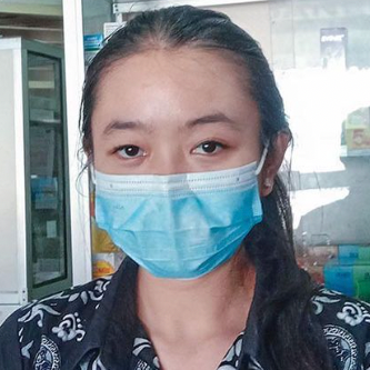 Seorang wanita mengenakan masker wajah biru di dalam, menatap kamera. Ia berada di depan lemari kaca.
