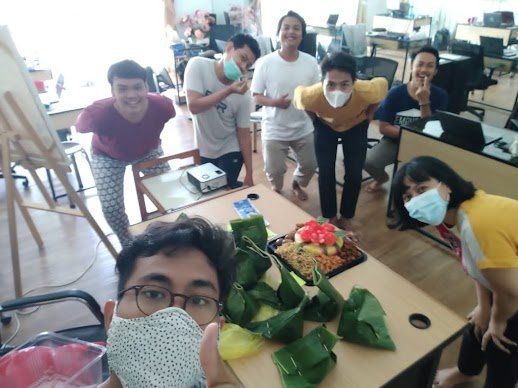 Sekelompok orang mengelilingi meja dengan makanan, beberapa mengenakan masker, tersenyum ke arah kamera. Suasana di dalam ruangan.