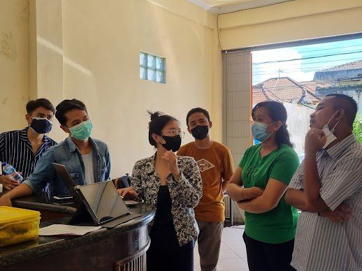Sekelompok orang bermasker sedang menatap tablet di atas meja, mungkin sedang berdiskusi. Di dalam ruangan, pencahayaan terang.