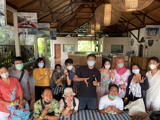 Sekelompok orang bertopeng berpose di dalam ruangan, beberapa di antaranya memasang tanda perdamaian. Atap kayu, warna netral.