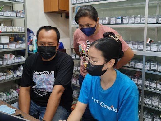 Tiga orang mengenakan masker sedang menatap komputer di apotek dengan rak-rak obat.