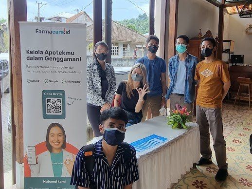 Sekelompok orang berpose dengan spanduk Farmacare.id di dalam ruangan. Beberapa mengenakan masker. Spanduk tersebut memiliki kode QR dan wajah seorang perempuan.