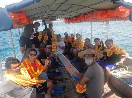 Orang-orang mengenakan rompi pelampung di atas perahu di laut, beberapa melambaikan tangan dan tersenyum. Air dan langit biru.