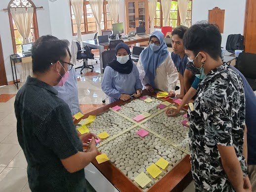 Orang-orang berkumpul di sekitar meja dengan catatan tempel. Di dalam ruangan. Cahaya alami.