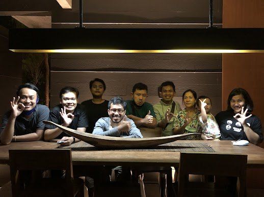 Sekelompok orang di meja kayu, tersenyum dan memberi isyarat, diterangi oleh lampu panjang.