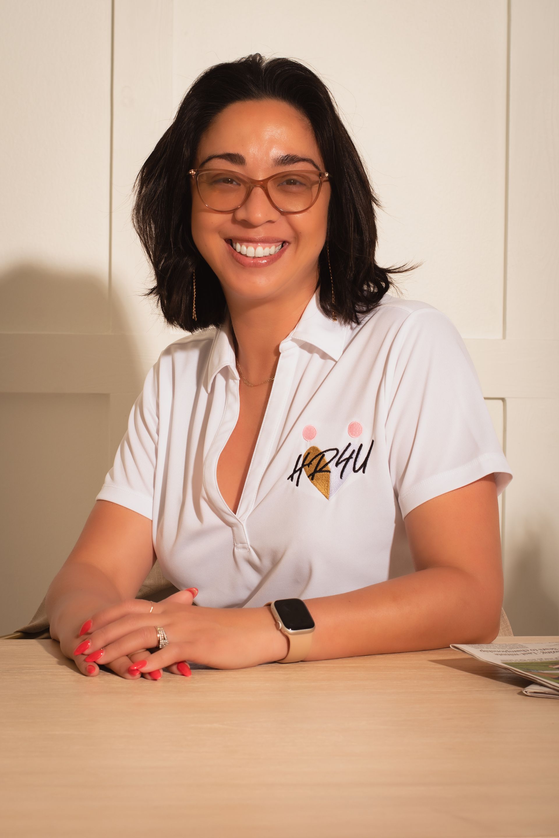 Woman with glasses smiles, wearing a white shirt with a pink logo. She sits at a light-colored table.
