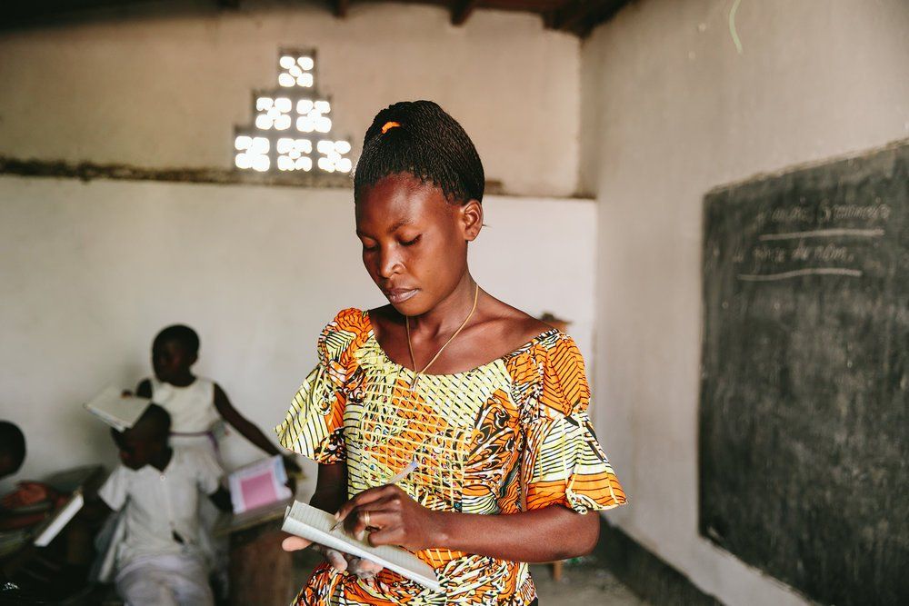 Girl in patterned dress writes in notebook in a classroom; students sit nearby.