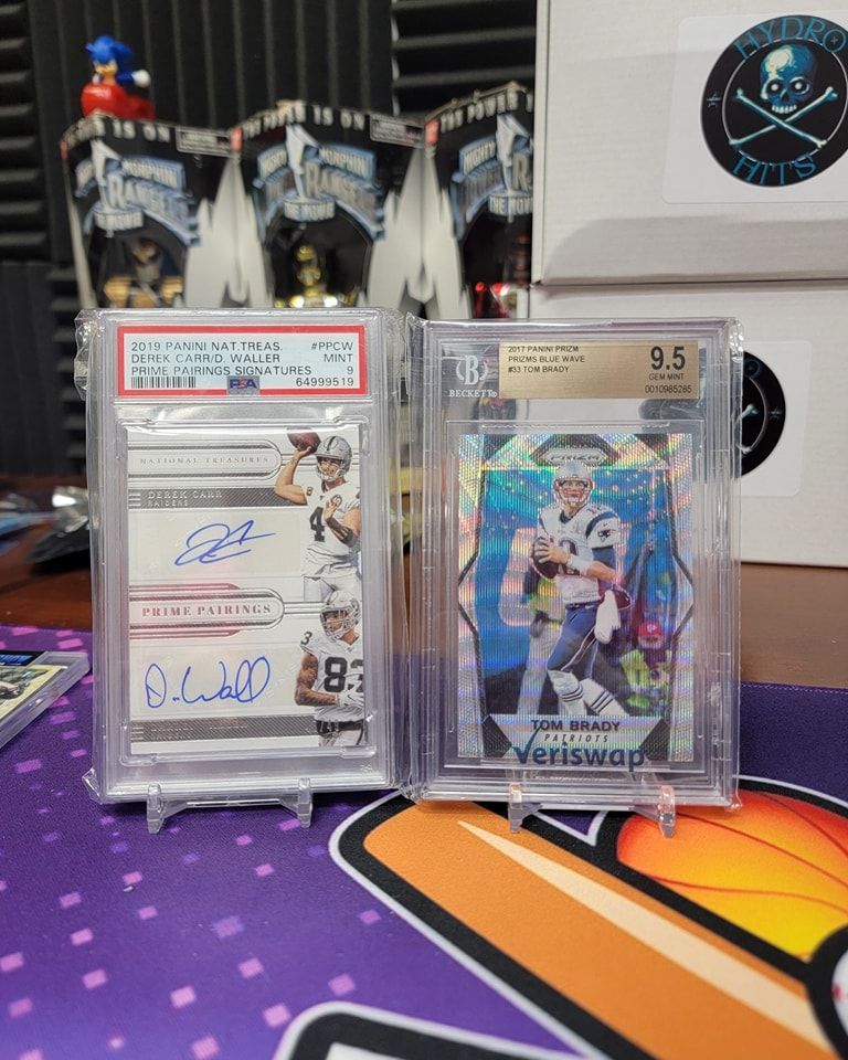 Two graded sports cards in clear stands on a colorful desk, with boxes and memorabilia behind them.