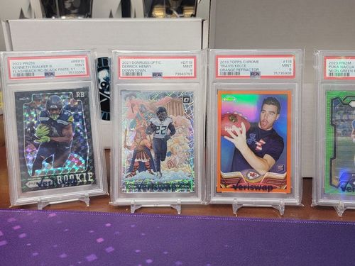 Graded trading cards in plastic slabs displayed on clear stands against a purple dotted surface.