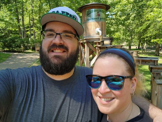 Two smiling people taking a selfie in a sunny wooded park near a water fountain
