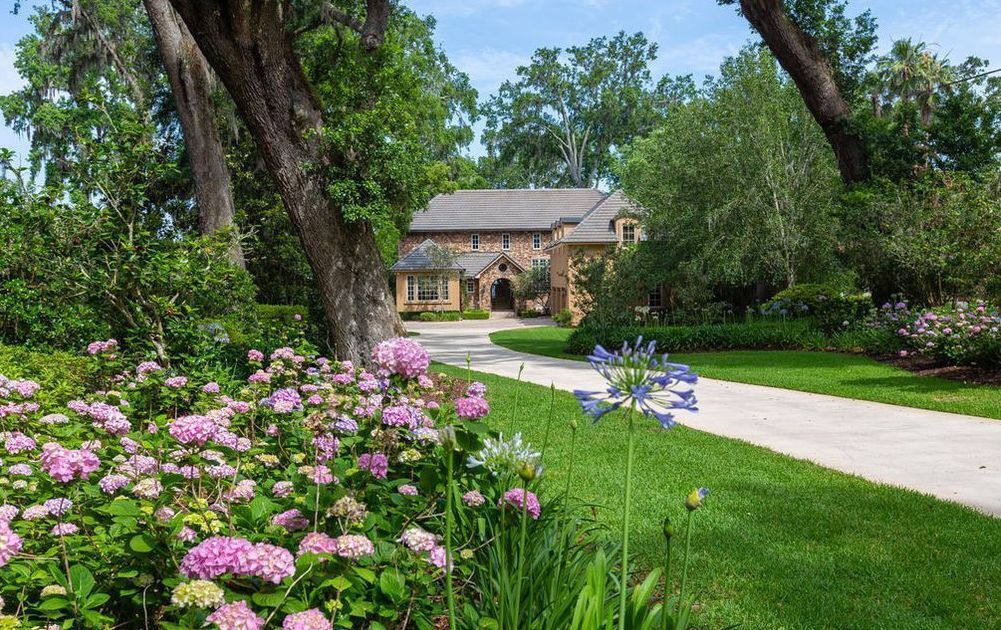 A house with a driveway leading to it is surrounded by flowers and trees.