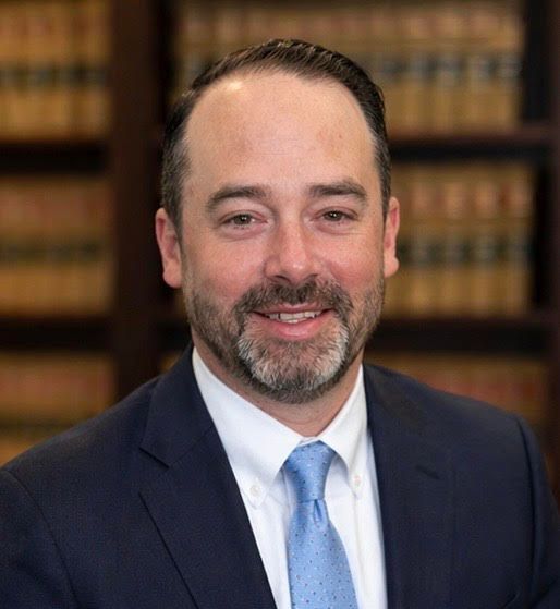 Smiling man in a navy suit and blue tie, posing in front of bookshelves.