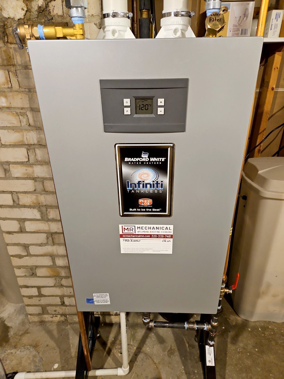 A water heater is sitting in a basement next to a brick wall.