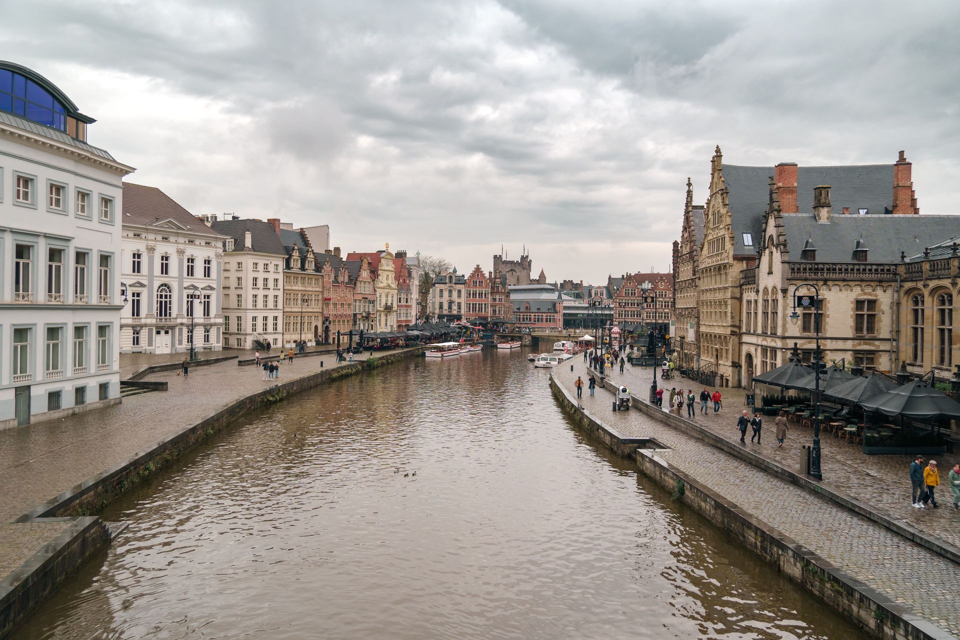 Canal lined with historic buildings under a cloudy sky, with people walking along the waterfront.