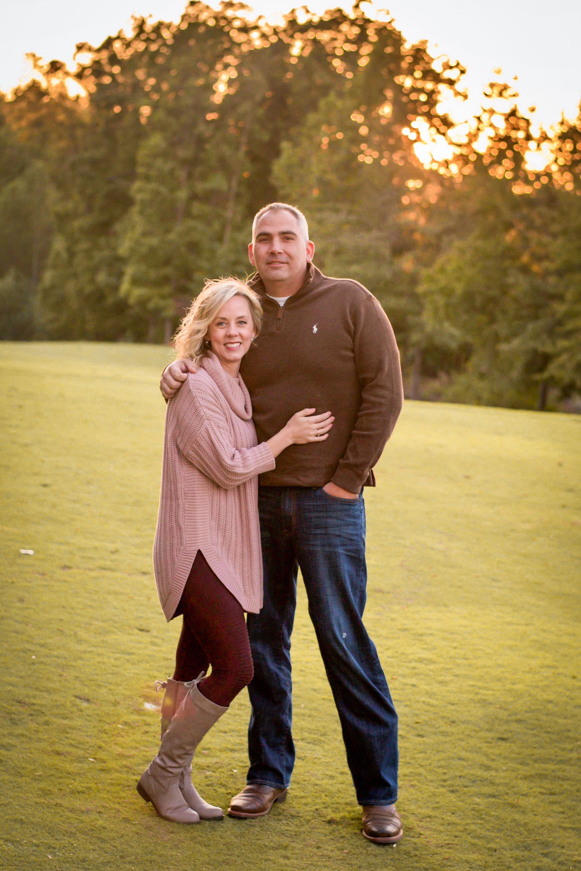 Couple embracing on a green lawn with trees in the background, sun setting.